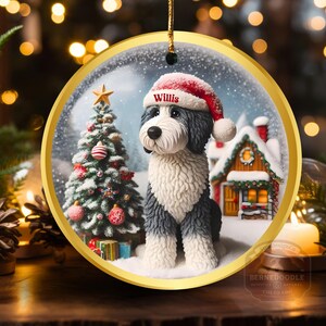 May include: A circular Christmas decoration with a gold edge depicts a dog in a Santa hat, labelled "Willis". The ornament shows a winter scene with a Christmas tree and a snow-covered house. It is suspended by a gold string.