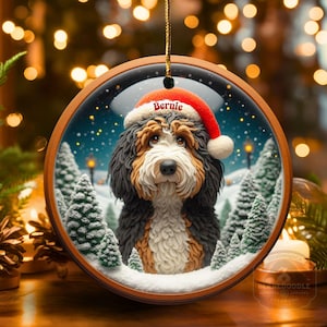 Op de afbeelding: Een ronde houten kerstversiering met een Bernedoodle hond met een kerstmuts met de naam "Bernie". De versiering heeft een winterse achtergrond met besneeuwde bomen. Het hangt aan een gouden touwtje.