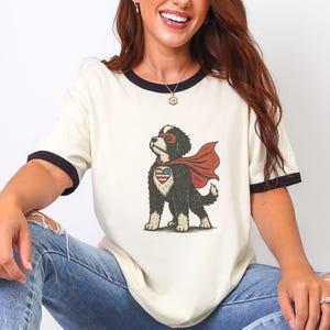 May include: Cream-coloured t-shirt with black trim featuring a cartoon dog superhero with a red cape and an American flag heart. The dog is black and white. The shirt is paired with blue jeans and white trainers.