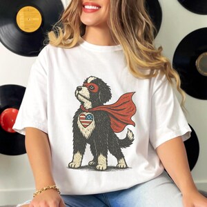 May include: A white short-sleeve t-shirt featuring a graphic of a black and white dog wearing a red superhero mask and cape. The dog has a heart-shaped patch on its chest with a red, white, and blue American flag pattern.
