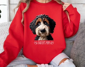 Peut inclure: Un sweat-shirt rouge avec une illustration détaillée du visage d'un chien Bernedoodle. Le texte "Baxter's Mom" est imprimé sous l'image du chien, avec des symboles de cœur de chaque côté. Le sweat-shirt est de couleur rouge uni.