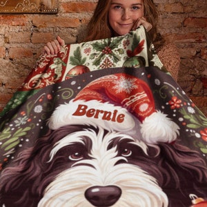 May include: A Christmas-themed blanket featuring a dog wearing a Santa hat with the name "Bernie". The blanket has a festive design with red and white ornaments and floral accents. The dog's face is the focal point, with a brown and white color scheme.