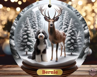 Könnte beinhalten: Runder Weihnachtsschmuck mit Schneekugel-Design. Er zeigt ein Reh und einen Hund in einer verschneiten Waldszene. Das Ornament hat ein goldenes Etikett mit dem Namen "Bernie". Aufgehängt an einer goldfarbenen Schnur.
