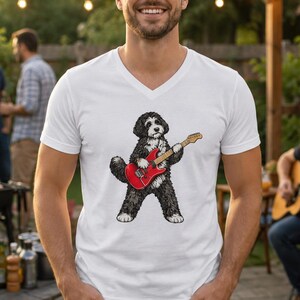 May include: A white V-neck t-shirt featuring a graphic illustration of a black and white dog standing on its hind legs while playing a bright red electric guitar. The dog has a fluffy coat and is positioned in the center of the shirt.