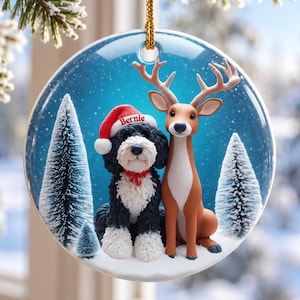 May include: A round ceramic Christmas ornament featuring a black and white dog wearing a red Santa hat with the name Bernie printed on it, sitting next to a brown deer in a snowy forest setting with pine trees and a blue background.