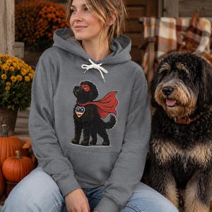 Pu&ograve; includere: Una felpa con cappuccio grigio melange con la grafica di un cane nero che indossa una maschera e un mantello da supereroe rossi, con un'etichetta a forma di cuore con motivo a stelle rosso, bianco e blu sul petto e cordini bianchi.