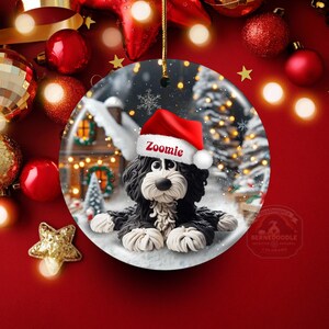 May include: A round Christmas ornament featuring a black and white Bernedoodle dog wearing a red Santa hat with the name "Zoomie". The ornament has a winter scene background with a house, snow, and Christmas lights. The ornament is hanging with a gold string.