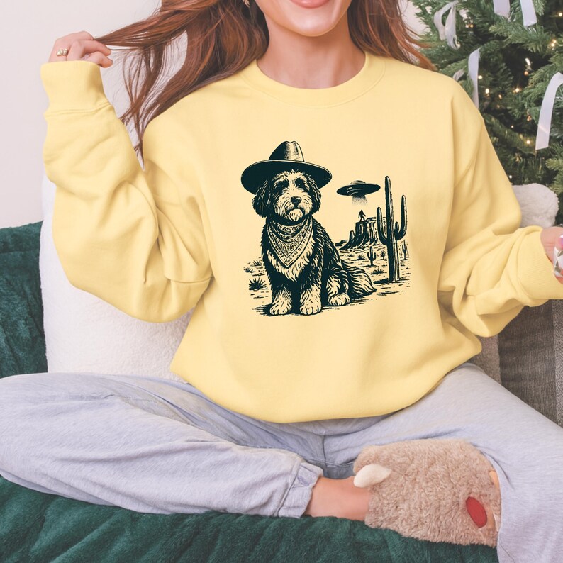 May include: A light yellow sweatshirt featuring a black and white illustration of a dog wearing a cowboy hat and bandana, with a desert landscape and UFOs in the background. The sweatshirt is a casual style.