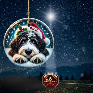 May include: A round Christmas ornament featuring a Bernedoodle dog wearing a Santa hat with the name "Bernie" on it. The ornament has a gold string for hanging and is set against a snowy, starry background.