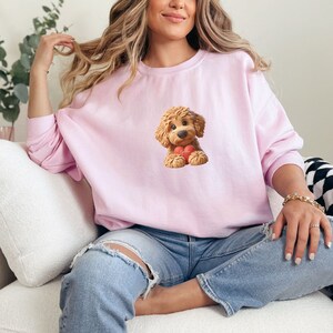 May include: A light pink sweatshirt featuring a cartoon illustration of a fluffy, brown dog holding a red heart. The dog has a sweet expression. The sweatshirt is a solid color and has long sleeves.