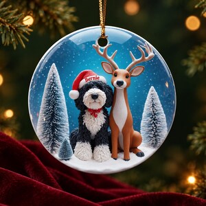 May include: A round ceramic Christmas ornament featuring a black and white dog wearing a red Santa hat with the name Bernie printed on it, sitting next to a brown deer. The scene includes snowy ground and small pine trees against a blue background.