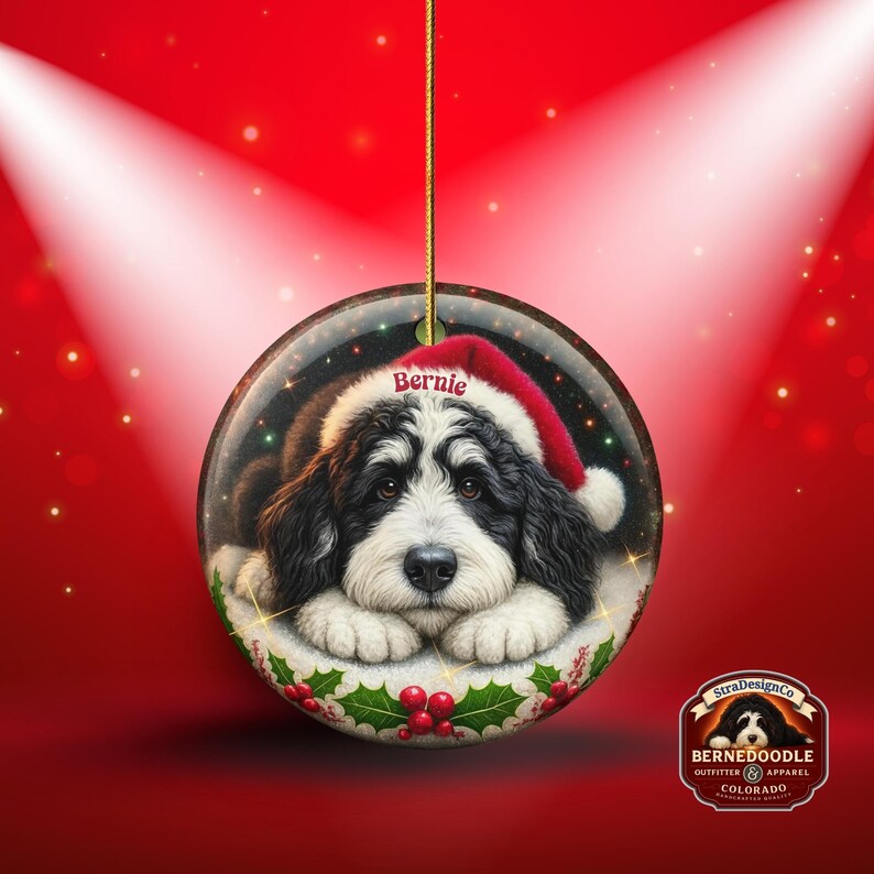 May include: A round Christmas ornament featuring a Bernedoodle dog wearing a Santa hat with the name "Bernie". The ornament has a red and white holly berry design around the base and is suspended by a gold string. The background is red.