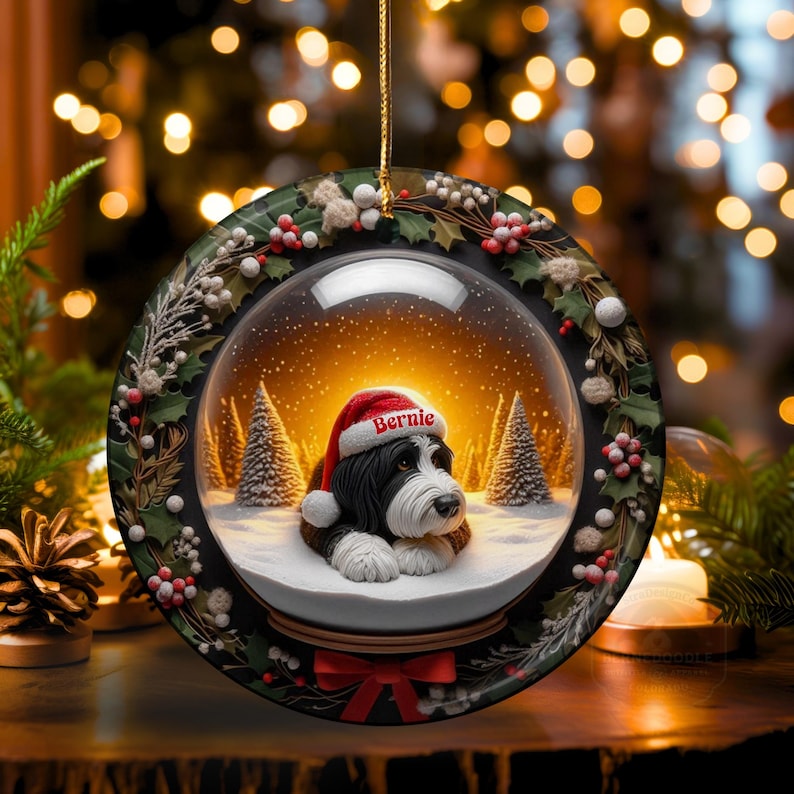 May include: A round Christmas ornament featuring a snow globe design. Inside, a dog wearing a Santa hat with the name "Bernie" is nestled in a snowy scene with pine trees. The ornament is trimmed with holly and berries.