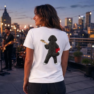 May include: A white short-sleeved t-shirt featuring a graphic on the back of a black curly-haired dog standing on its hind legs, holding a light-colored electric guitar and a red heart shape. The dog has white paws and a textured, shaggy appearance.