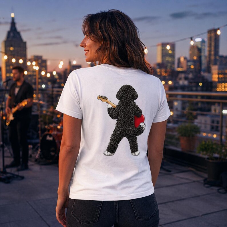 May include: A white short-sleeved t-shirt featuring a graphic on the back of a black curly-haired dog standing on its hind legs, holding a light-colored electric guitar and a red heart shape. The dog has white paws and a textured, shaggy appearance.