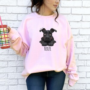 Puede incluir: Sudadera rosa claro con un dibujo de cachorro negro y el nombre "KENZIE" debajo. El cachorro tiene ojos grandes y expresivos. La sudadera es de color liso y tiene mangas largas.