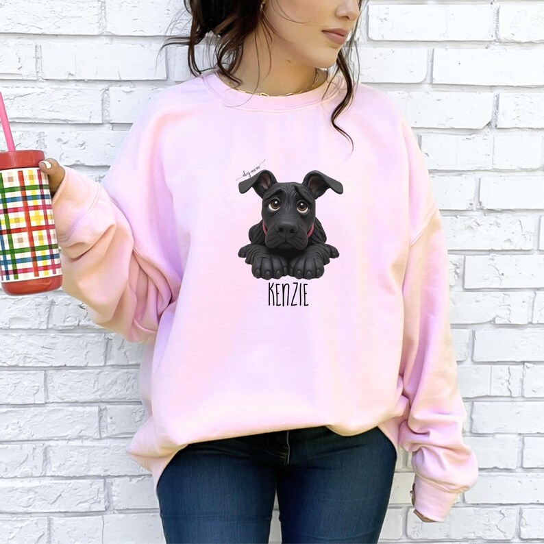 Puede incluir: Sudadera rosa claro con un dibujo de cachorro negro y el nombre "KENZIE" debajo. El cachorro tiene ojos grandes y expresivos. La sudadera es de color liso y tiene mangas largas.