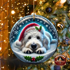 May include: Round Christmas ornament featuring a white dog wearing a red Santa hat with the word "snowflake". The ornament has a snowy background with holly and berries. The ornament is hanging in front of a Christmas tree.