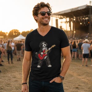May include: A black V-neck t-shirt featuring a graphic illustration of a fluffy black and white dog playing a bright red electric guitar. The dog has a shaggy coat and is standing on its hind legs while holding the guitar with its front paws.