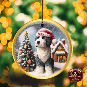 May include: A circular Christmas decoration with a Bernedoodle dog wearing a Santa hat with the name "Willis". The ornament has a gold border and shows a snowy scene with a Christmas tree and a house. The ornament is from StraDesignCo.