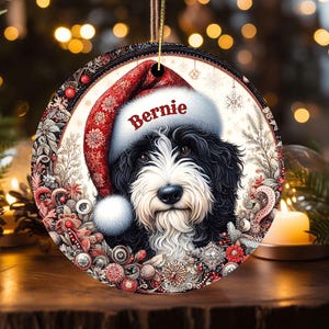 May include: Round Christmas ornament featuring a black and white dog wearing a Santa hat with the name "Bernie" in red. The ornament has a decorative border with red and white floral elements and a gold string for hanging.