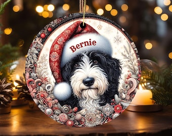 May include: Round Christmas ornament featuring a black and white dog wearing a Santa hat with the name "Bernie" in red. The ornament has a decorative border with red and white floral elements and a gold string for hanging.