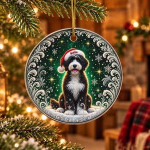 May include: A round ceramic Christmas ornament featuring a black and white dog wearing a red Santa hat with the name Stella printed on the brim. The dog sits in snow against a dark green background with white snowflakes and silver filigree.