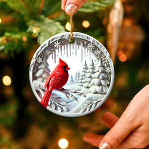 Peut inclure: Ornement de No&euml;l rond et blanc, orn&eacute; d'une sc&egrave;ne hivernale d&eacute;taill&eacute;e. Un cardinal rouge vif est perch&eacute; sur une branche enneig&eacute;e, entour&eacute; de sapins blancs et de flocons de neige. L'ornement est suspendu par une ficelle dor&eacute;e.