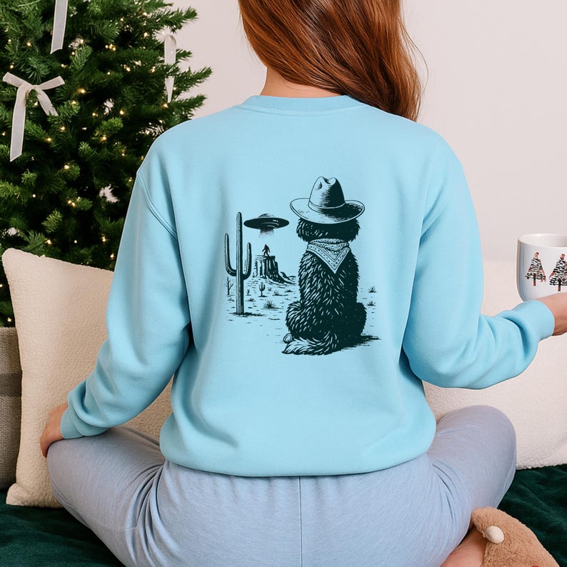 May include: Light blue sweatshirt featuring a graphic of a dog wearing a cowboy hat and bandana, set against a desert landscape with a UFO. The design is in black and white. The sweatshirt is a solid light blue color.
