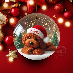 May include: A clear glass Christmas bauble featuring a brown dog wearing a Santa hat with the name "Chippy" on it. The dog is lying in snow with small Christmas trees in the background. The bauble is suspended against a red background with Christmas decorations.