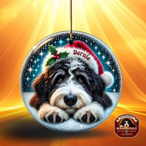 May include: A round Christmas ornament featuring a Bernedoodle dog wearing a Santa hat with the name "Bernie". The ornament has a snowy background with holly and berries. The ornament is hanging from a gold string.