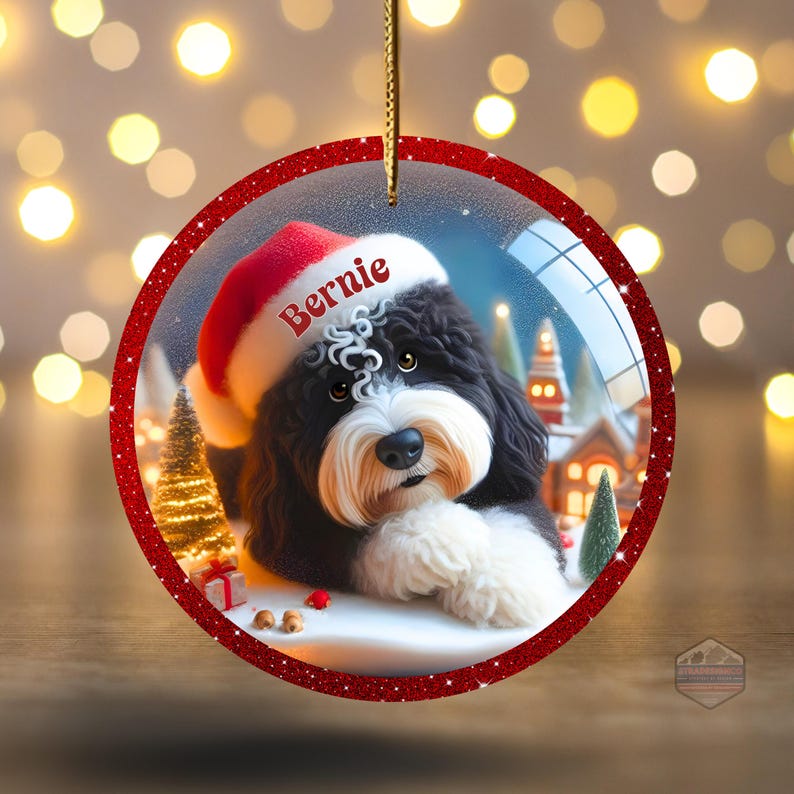 K&ouml;nnte beinhalten: Runder Weihnachtsschmuck mit einem schwarz-wei&szlig;en Hund, der einen Weihnachtsmannhut mit dem Namen "Bernie" tr&auml;gt. Der Schmuck hat einen roten Glitzerrahmen und eine goldene Schnur zum Aufh&auml;ngen. Der Hintergrund zeigt eine Winterlandschaft mit Schnee und einem Weihnachtsbaum.
