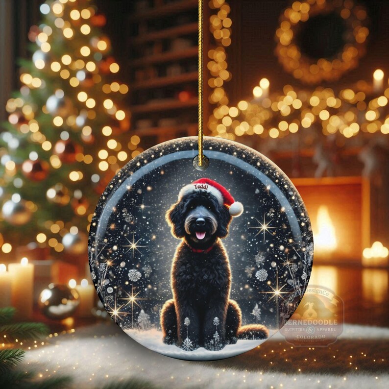 May include: Round Christmas ornament featuring a black dog wearing a Santa hat with the name "Teddy". The ornament has a snowy winter scene with a Christmas tree and a fireplace in the background. The ornament is hanging by a gold string.