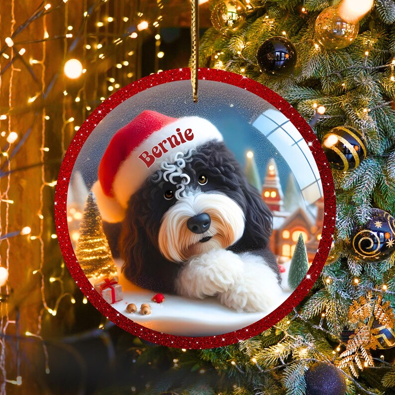 K&ouml;nnte beinhalten: Ein runder Weihnachtsschmuck mit einem schwarz-wei&szlig;en Hund, der einen Weihnachtsmannhut tr&auml;gt und den Namen "Bernie" tr&auml;gt. Der Schmuck hat einen roten Glitzerrahmen und eine goldene Schnur zum Aufh&auml;ngen. Der Hintergrund zeigt eine verschneite Szene mit H&auml;usern und Weihnachtsb&auml;umen.