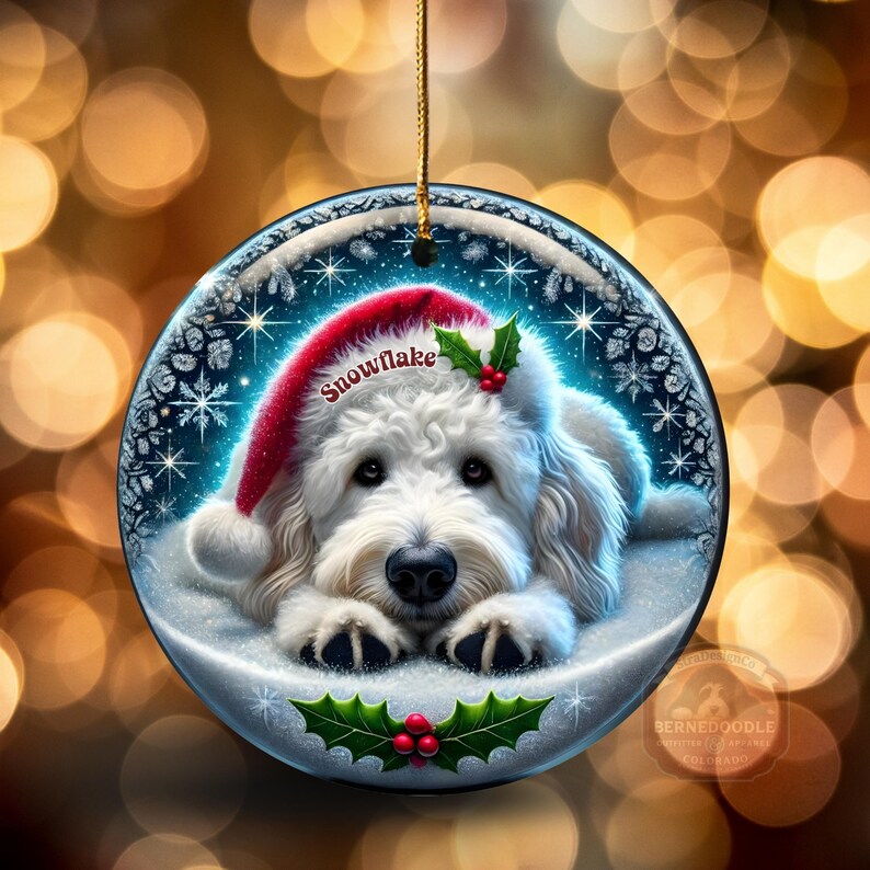 May include: A round Christmas ornament featuring a white dog wearing a red Santa hat with the word "Snowflake". The ornament has a gold string for hanging and is decorated with holly and snowflakes.