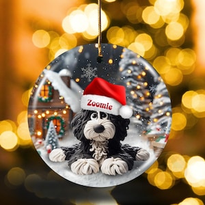 May include: A round Christmas ornament featuring a black and white dog wearing a red Santa hat with the name "Zoomie". The ornament has a winter scene background with a snowy house and trees.