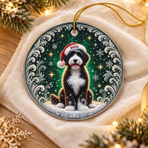 May include: A round ceramic ornament featuring a black and white dog wearing a red Santa hat with the name Stella printed on the brim. The dog sits against a dark green background with white snowflakes, gold stars, and ornate silver scrollwork.