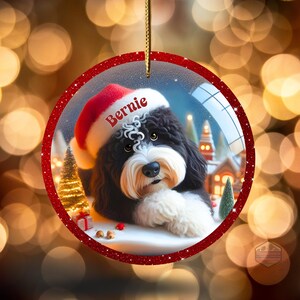 K&ouml;nnte beinhalten: Runder Weihnachtsschmuck mit einem schwarz-wei&szlig;en Hund mit Weihnachtsm&uuml;tze und dem Namen "Bernie". Der Schmuck hat einen roten Glitzerrahmen und eine goldene Schnur zum Aufh&auml;ngen. Der Hintergrund zeigt eine Winterlandschaft.