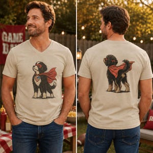 May include: A beige V-neck t-shirt featuring a graphic of a black and tan dog wearing a red superhero cape and a mask. The front graphic shows the dog with a heart-shaped American flag emblem, while the back graphic shows the dog from behind.