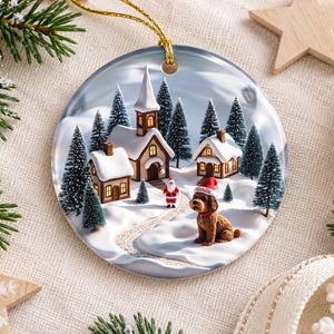 May include: A round ceramic Christmas ornament featuring a snowy village scene with a church, houses, pine trees, a small Santa Claus figure, and a brown dog wearing a red hat with the name Goldie printed on it, suspended by a gold cord.