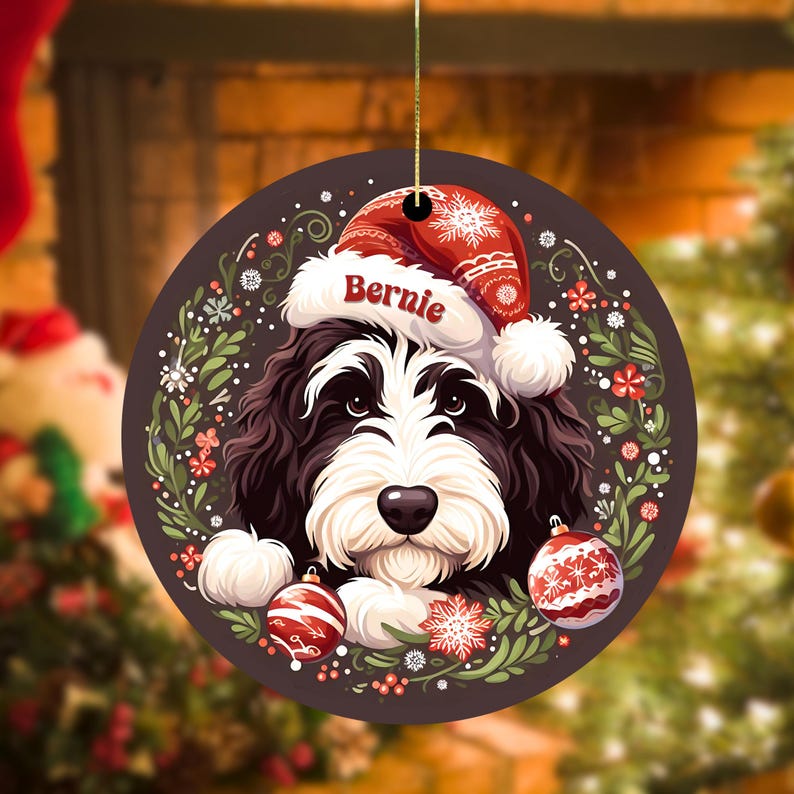 May include: A round Christmas ornament featuring a black and white dog wearing a red Santa hat with the name "Bernie". The ornament is decorated with red and white ornaments, snowflakes, and greenery. A gold string is attached for hanging.