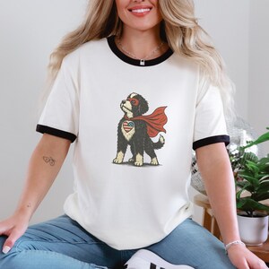 May include: Cream-coloured ringer t-shirt with black trim featuring a cartoon dog superhero. The dog has a red cape, mask, and a heart-shaped emblem with an American flag design. The shirt is worn by a person.