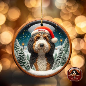 Op de afbeelding: Een ronde kerstversiering met een Bernedoodle-hond met een kerstmuts met de naam "Bernie". De versiering heeft een winterse achtergrond met besneeuwde bomen en een houten frame. De versiering is van StraDesignCo.