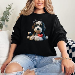 A woman wearing a Bernedoodle sweatshirt with cute design on the front and the back.  The front features a bernedoodle dog in claymation design holding a heart with his name, across the top of his head the words, "Doodle Mom".