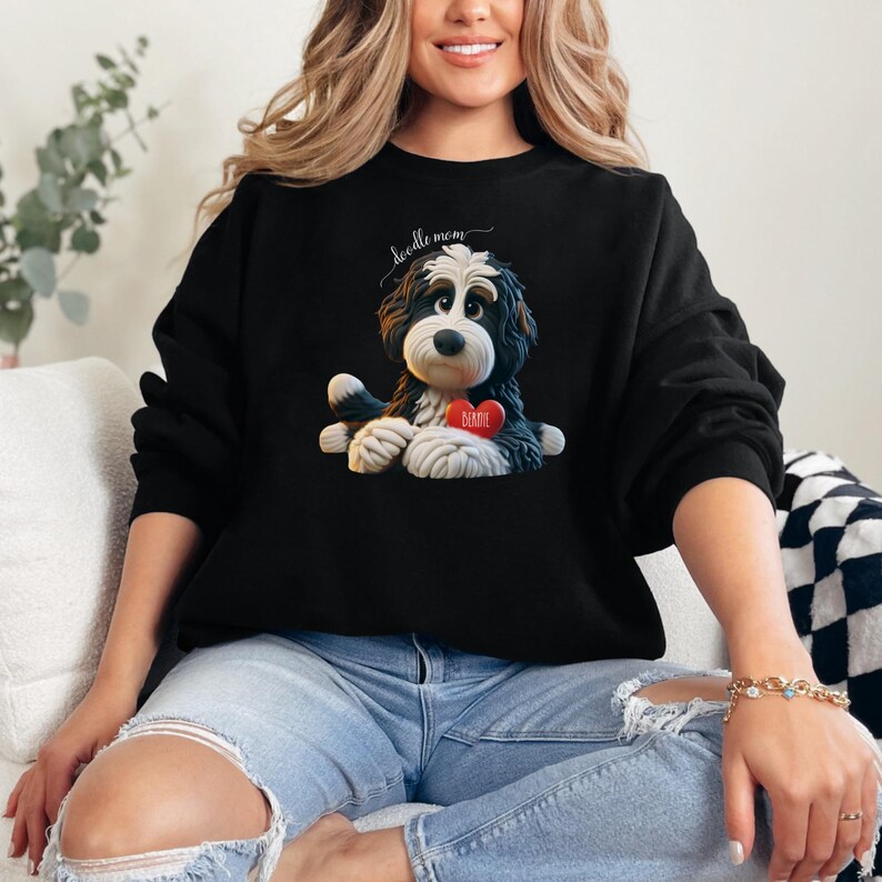A woman wearing a Bernedoodle sweatshirt with cute design on the front and the back.  The front features a bernedoodle dog in claymation design holding a heart with his name, across the top of his head the words, "Doodle Mom".