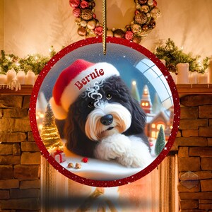 K&ouml;nnte beinhalten: Ein runder Weihnachtsschmuck mit einem schwarz-wei&szlig;en Hund, der einen Weihnachtsmannhut mit dem Namen "Bernie" tr&auml;gt. Der Schmuck hat einen roten Glitzerrahmen und eine goldene Schnur zum Aufh&auml;ngen. Der Hintergrund zeigt eine Winterlandschaft.
