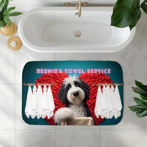 May include: A rectangular bath mat featuring a cartoon dog with black and white fur, a red heart-shaped design, and the text "BERNIE'S TOWEL SERVICE" in pink. White towels hang on a clothesline. The mat is placed in front of a white bathtub.