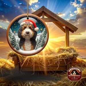 Op de afbeelding: Een ronde kerstversiering met een Bernedoodle-hond met een kerstmuts met de naam "Bernie". De versiering heeft een winterse achtergrond met besneeuwde bomen. Het hangt aan een gouden touwtje.