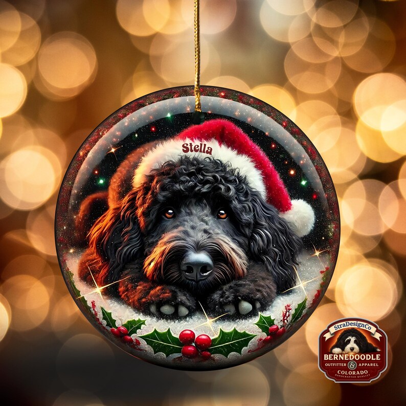 May include: Round Christmas ornament featuring a black dog wearing a Santa hat. The dog's name, "Stella," is printed on the hat. The ornament has a gold string for hanging and is decorated with holly and berries.