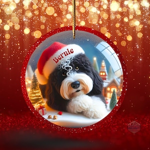 K&ouml;nnte beinhalten: Ein runder Weihnachtsschmuck mit einem schwarz-wei&szlig;en Hund, der einen Weihnachtsmannhut mit dem Namen "Bernie" tr&auml;gt. Der Schmuck hat einen roten Rand und einen goldenen Faden zum Aufh&auml;ngen. Der Hintergrund zeigt eine verschneite Szene.
